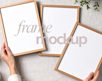 May include: Three wooden picture frames with white inserts are arranged on a light-colored surface. The frames are held by hands, and the text "frame mockup" is superimposed on the frames. The frames have a natural wood finish.