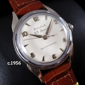 Puede incluir: Un reloj de pulsera Bulova 23 Jewel de cuerda automática con una correa de cuero marrón. La esfera es blanca con agujas e indicadores de hora plateados. El texto "Bulova 23 Jewel SELF WINDING" es visible. El reloj es de c.1956.