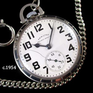 May include: A vintage Hamilton Railway Special pocket watch with a white face, black numerals, and a subdial. The watch has a silver-tone case and chain. The text "HAMILTON RAILWAY SPEC" is visible. The image also includes the text "c.1954".
