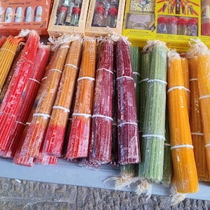 Pode incluir: Feixes de velas cônicas coloridas em tons de amarelo, vermelho, laranja, verde e bordô. As velas são embaladas em plástico transparente e amarradas com barbante branco. Várias caixas de óleo de unção e especiarias são visíveis.