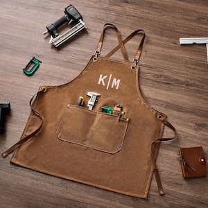 May include: A brown canvas apron with cross-back leather straps and the white embroidered letters K|M on the chest. The apron features a large front pocket containing a caliper and small tools, surrounded by a nail gun, a square, and a small notebook.