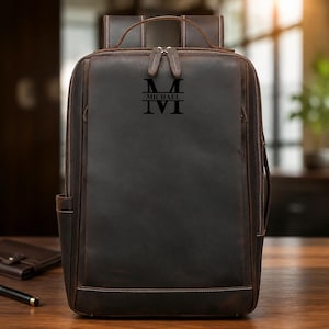 May include: A dark brown leather backpack featuring a rectangular silhouette, a top handle, and two shoulder straps. The front panel is engraved with a large letter M, the name MICHAEL, and the letter M below it in a stacked monogram design.