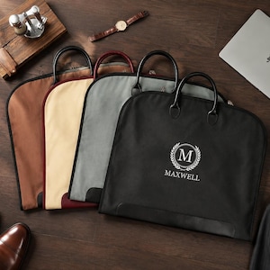 May include: Four garment bags in black, grey, beige, and brown, each featuring two black handles and a zipper closure. The black bag displays a white circular logo with the letter M inside a laurel wreath and the text MAXWELL printed below.