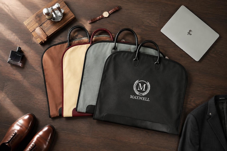 May include: Four garment bags in black, grey, beige, and brown, each featuring two black handles and a zipper closure. The black bag displays a white circular logo with the letter M inside a laurel wreath and the text MAXWELL printed below.