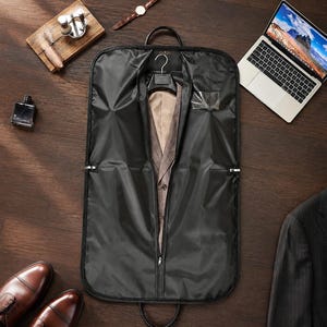 May include: A black garment bag with a central zipper opening, revealing a tan suit jacket on a hanger inside. The bag features a clear plastic window for identification and two top handles for carrying, designed to protect clothing during travel.