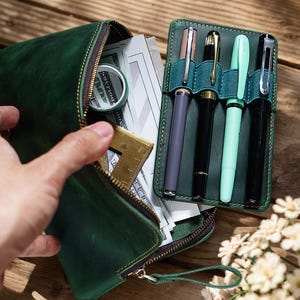 Puede incluir: Una bolsa de cuero verde con cremallera y un portaplumas. La bolsa está abierta y contiene bolígrafos, una regla y un rollo de cinta.