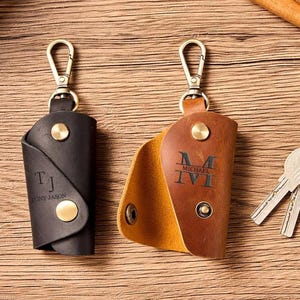 May include: Two leather key holders with metal clasps and gold-coloured hardware. One is black with the initials "TJ" and the name "TONY JASON" engraved. The other is brown with the initials "M" and "IVI" engraved.
