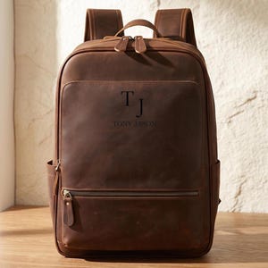 May include: A brown leather backpack featuring a top handle, two shoulder straps, and a front zippered pocket. The front panel is engraved with the initials TJ and the name TONY JASON. The bag has a smooth, matte finish with visible stitching.