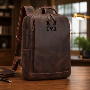 Op de afbeelding: Een donkerbruine leren rugzak met een rechthoekig silhouet, verstelbare schouderbanden en een handvat aan de bovenkant. Het voorpaneel is gepersonaliseerd met een zwart monogramontwerp bestaande uit een grote letter M en de naam MICHAEL.