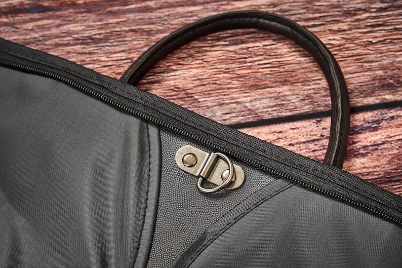 May include: A black bag featuring a smooth fabric exterior with a textured central panel, a dark brown leather-like handle, and a metallic D-ring clasp secured with two rivets. The bag includes a black zipper closure along the top edge.