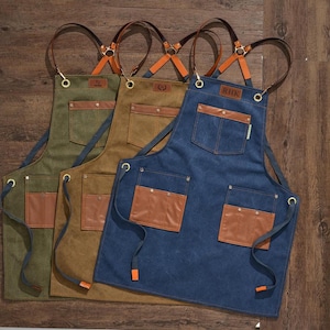 May include: Three canvas aprons with leather accents and adjustable straps. The aprons are in different colours: green, brown, and blue. The blue apron has the initials "RHK" on the front.