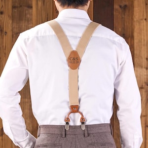 May include: Tan suspenders with brown leather accents and silver-tone clips. The leather patch features the initials "C&S" and the date "10.22.20". The suspenders are worn over a white dress shirt and brown trousers, against a wooden background.
