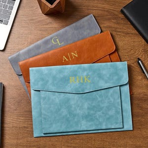 Peut inclure: Trois pochettes de documents en cuir personnalisées, grises, orange et turquoise, chacune avec des initiales dorées. La pochette turquoise est au premier plan, avec les initiales "RHK". Un ordinateur portable, un stylo et un carnet sont également visibles.