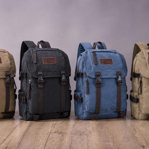 Può includere: Quattro zaini in tela disposti in fila, dotati di una patta superiore con toppe in similpelle marrone, cinghie in tessuto marrone scuro e chiusure con fibbia in plastica. Gli zaini sono disponibili nei colori beige, nero, blu e beige chiaro.