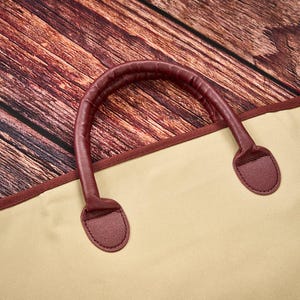 May include: A beige garment bag featuring a dark maroon trim and a matching dark maroon rounded handle. The bag is constructed with a smooth, solid-colored fabric and includes reinforced stitching where the handle attaches to the main body.