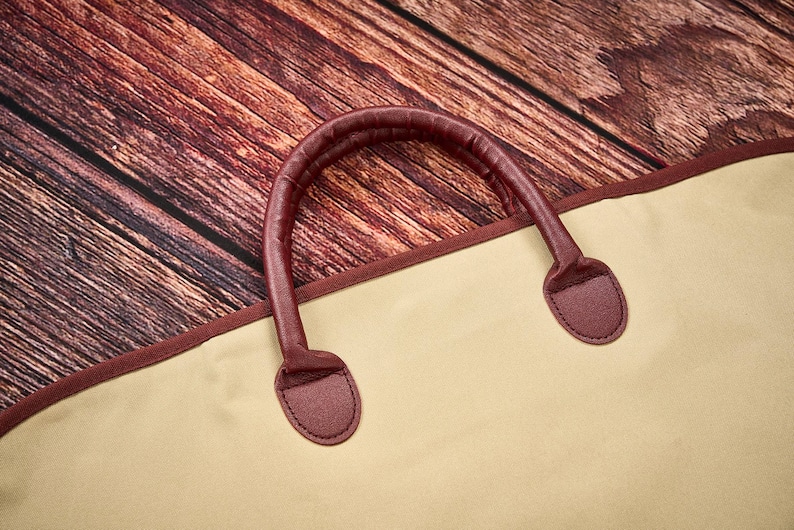 May include: A beige garment bag featuring a dark maroon trim and a matching dark maroon rounded handle. The bag is constructed with a smooth, solid-colored fabric and includes reinforced stitching where the handle attaches to the main body.