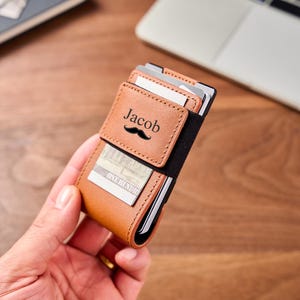 May include: A tan leather card holder wallet featuring a black elastic band and a metal frame. The front of the wallet is engraved with the name Jacob above a black moustache icon. Several credit cards and a one hundred dollar bill are visible.