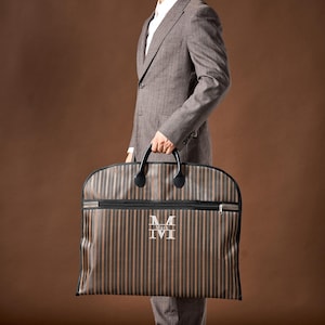 May include: A brown and black striped garment bag featuring a horizontal zipper across the top and a central white monogram design with the name Mark printed inside the letter M. The bag includes two black handles attached to the top edge.