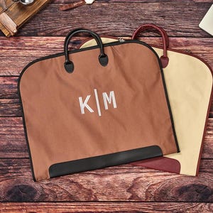 May include: A brown garment bag with black handles and a black bottom trim, featuring the white embroidered letters K|M in the center. Behind it, a second garment bag in cream with a maroon handle and trim is partially visible.