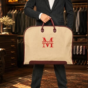 Puede incluir: Una funda para ropa de color beige con ribetes y asas de color rojo oscuro. El centro presenta una gran letra M bordada en rojo con el nombre MICHAEL impreso horizontalmente en medio de la letra. La bolsa tiene una parte superior redondeada.