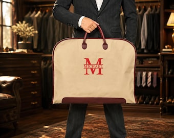 May include: A beige garment bag with dark red trim and handles. The centre features a large red embroidered letter M with the name MICHAEL printed horizontally across the middle of the letter. The bag has a rounded top and a rectangular base.