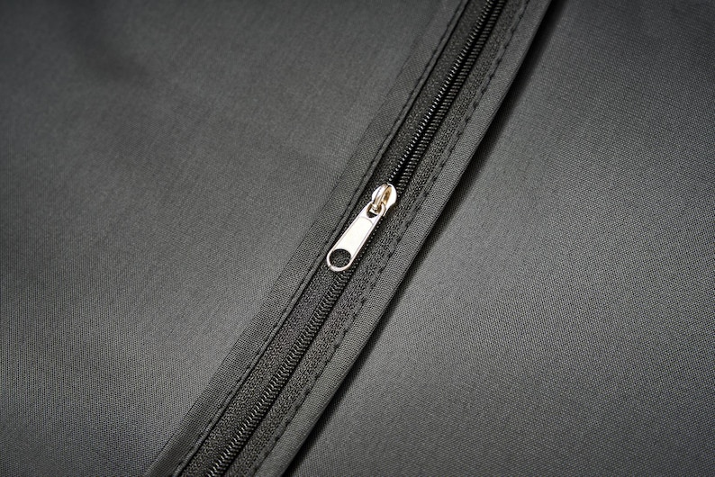 May include: A close-up view of a dark gray fabric surface featuring a central black zipper closure. The zipper track is integrated into the material with a silver-toned metal pull tab positioned vertically along the center of the textile panel.
