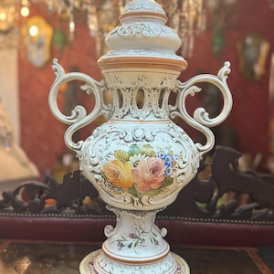 Puede incluir: Un jarrón de cerámica grande y ornamentado, de color crema, con tapa, asas y diseño floral. El jarrón presenta un arreglo floral detallado, pintado a mano, en tonos amarillos, rosas y azules. El jarrón mide aproximadamente 61 cm de alto y se asienta sobre una base decorativa.
