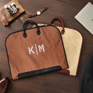 May include: Two garment bags with handles, one in brown and one in beige. The brown bag features the white embroidered letters K|M. Both bags have black trim and zippers, designed for carrying clothing items while maintaining a structured shape.