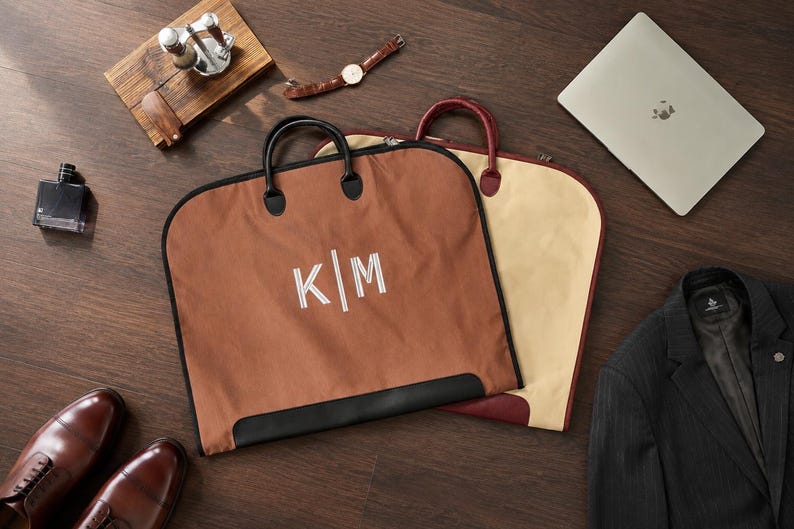 May include: Two garment bags with handles, one in brown and one in beige. The brown bag features the white embroidered letters K|M. Both bags have black trim and zippers, designed for carrying clothing items while maintaining a structured shape.
