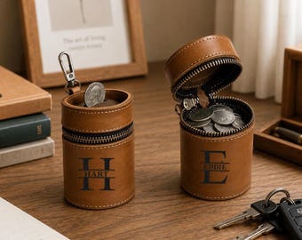 Peut inclure: Deux porte-monnaie cylindriques en cuir beige avec des fermetures éclair noires et des accessoires en métal. L'un est fermé par un mousqueton argenté, l'autre est ouvert et rempli de pièces. Chaque étui porte un monogramme avec les noms Hart et Eddie.
