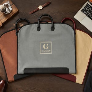 May include: Three garment bags in gray, tan, and burgundy, each featuring black handles and trim. The central gray bag displays a square emblem with the letter G and the name GARCIA printed in white. The bags are arranged on a dark wooden surface.