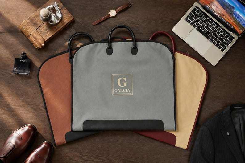 May include: Three garment bags in gray, tan, and burgundy, each featuring black handles and trim. The central gray bag displays a square emblem with the letter G and the name GARCIA printed in white. The bags are arranged on a dark wooden surface.