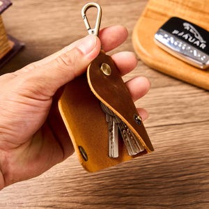 Puede incluir: Un llavero de cuero marrón con un cierre metálico, que contiene varias llaves. El llavero está abierto, revelando las llaves en su interior. Una llave de coche con el logotipo de Jaguar es visible en el fondo.