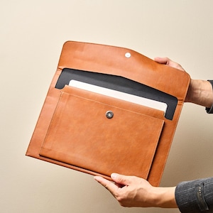 Könnte beinhalten: Eine braune Laptop-Hülle aus Leder mit Magnetverschluss. Die Hülle hat eine Vordertasche und ein schwarzes Innenfutter. Die Laptoptasche wird offen gehalten und zeigt das Innere und einen Laptop.