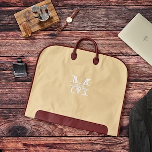 May include: A beige garment bag with dark red trim and handles, featuring a white embroidered monogram with the letter M and the name Mark centered on the front. The bag is rectangular with rounded corners and a solid dark red base panel.