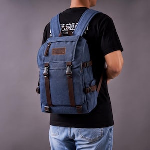 Puede incluir: Una mochila de lona azul con correas de aspecto de cuero marrón, hebillas de plástico negro y un parche rectangular marrón en la solapa superior. La mochila incluye correas para los hombros ajustables y correas de compresión laterales a juego.