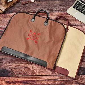May include: A brown garment bag with black trim and handles, featuring a red embroidered logo with crossed arrows and the letters W and M. A second garment bag in beige with a maroon trim is partially visible underneath the brown bag.