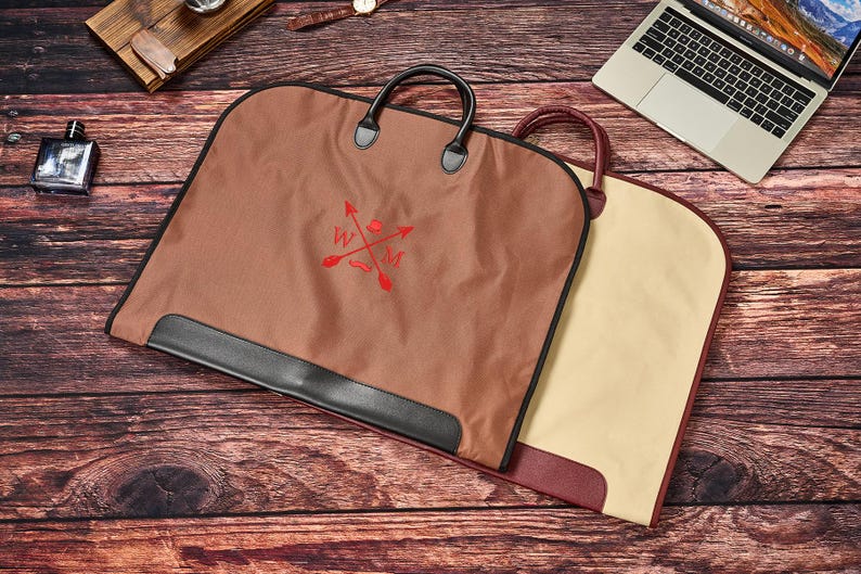 May include: A brown garment bag with black trim and handles, featuring a red embroidered logo with crossed arrows and the letters W and M. A second garment bag in beige with a maroon trim is partially visible underneath the brown bag.