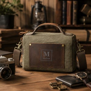 Op de afbeelding: Een olijfgroene canvas tas met een bruin lederen voorvak voorzien van de letter M en de naam Molina. De tas heeft een handvat aan de bovenkant, een afneembare bruine schouderriem met metalen clips en een ritssluiting aan de bovenzijde.