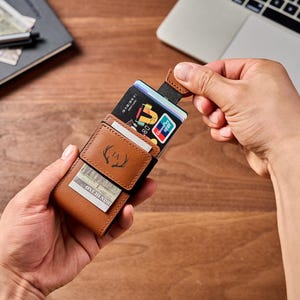 May include: A tan leather card holder wallet featuring a small embossed deer antler logo and the initials JA. The wallet has a pull-tab mechanism that partially extends a stack of credit cards, and a front exterior pocket containing a paper bill.