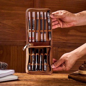 May include: A brown leather pen case, open to display several pens. The case is held open by a person's hands. The pens are black and silver, with clear ink cartridges. The case is on a wooden surface.