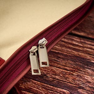 May include: A maroon bag featuring a dual zipper closure with two silver-toned metal pull tabs. The bag has a beige interior lining and a maroon exterior trim. The zipper track is also maroon, matching the exterior color of the bag's trim.