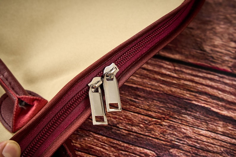 May include: A maroon bag featuring a dual zipper closure with two silver-toned metal pull tabs. The bag has a beige interior lining and a maroon exterior trim. The zipper track is also maroon, matching the exterior color of the bag's trim.