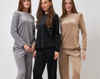 May include: Three women wearing matching velvet pajama sets in gray, black, and tan. Each set includes a long-sleeved top and wide-leg pants. The women are also wearing matching fuzzy slippers in coordinating colors.