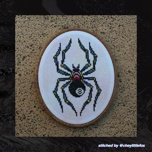 Puede incluir: Arte de punto de cruz ovalado con una ara&ntilde;a negra con un dise&ntilde;o de bola ocho en el abdomen, con acentos verdes y amarillos. La obra de arte est&aacute; enmarcada en un marco de madera sobre un fondo texturizado. El texto en la parte inferior dice "stitched by @cheylittlefox."