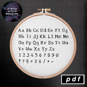 May include: A white embroidery hoop with a wooden frame. The hoop contains white Aida cloth with black cross-stitch lettering of the alphabet, numbers, and punctuation marks. The text reads: "Aa Bb Cc Dd Ee Ff Gg Hh Ii Jj Kk Ll Mm Nn Oo Pp Qq Rr Ss Tt Uu Vv Ww Xx Yy Zz 1234567890 !?@#%&/+- ". The text is in a typewriter font. The word "pdf" is printed in black on a white rectangle below the hoop.
