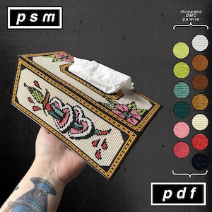 Puede incluir: Caja de pañuelos beige con un diseño de punto de cruz que presenta un corazón atravesado por una daga, flores y los textos "psm" y "pdf". La caja tiene un borde dorado y negro y contiene pañuelos blancos. Se muestra una paleta DMC enhebrada.