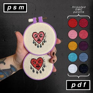 May include: Two cross-stitch embroidery hoops with a sad heart design. The hearts are red with black detailing and teardrops. The hoops are lavender, and the fabric is beige. A color palette of thread colors is shown, along with the text "threaded DMC palette" and "pdf".