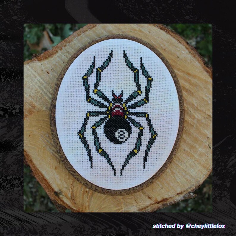 Puede incluir: Un dise&ntilde;o de ara&ntilde;a bordado a punto de cruz con cuerpo negro, motivo de bola ocho, patas verdes y detalles rojos. La obra est&aacute; en un marco de madera redondo, sobre una rodaja de madera. Texto: "stitched by @cheylittlefox".