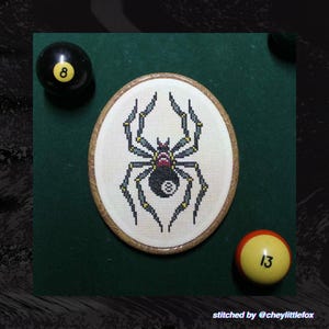 Puede incluir: Un bordado de punto de cruz ovalado con una ara&ntilde;a con un dise&ntilde;o de bola 8. La ara&ntilde;a es negra, gris y amarilla sobre un fondo blanco. Tambi&eacute;n hay dos bolas de billar, una negra con un 8 y otra amarilla con un 13.